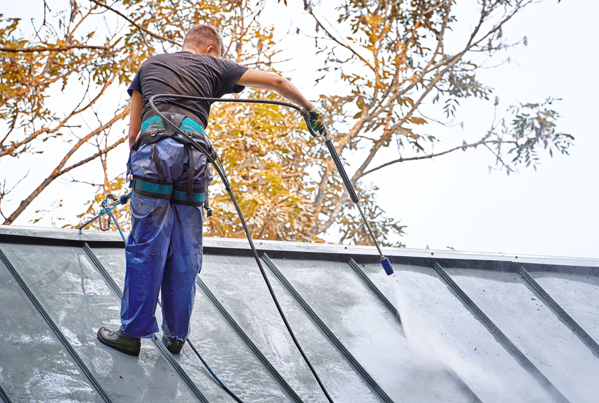 Worker in safety harness cleaning metal roof with pressure washer